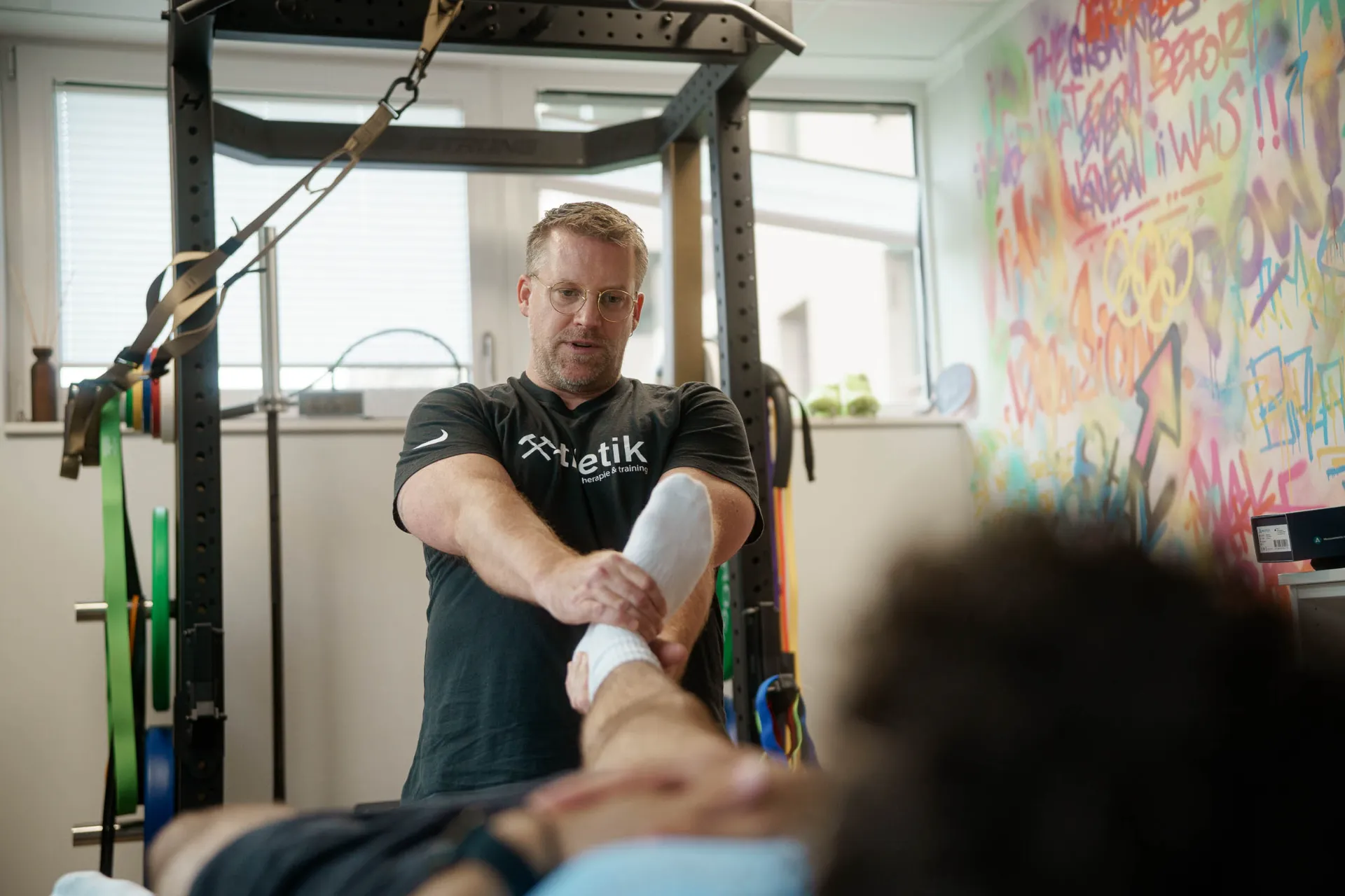 Sebastian Kaul, Inhaber von x-thletik, behandelt den Fuß eines Patienten in einem modernen Trainings- und Therapieraum mit bunter Graffiti-Wand – Teil der Darstellung des Behandlungskonzepts auf der Website.