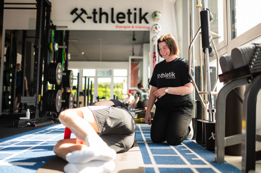 Eine x-thletik-Trainerin betreut einen Athleten, der auf einer blauen Trainingsmatte liegt und eine Mobilisationsübung ausführt. Das Bild unterstreicht, wie gezielte Betreuung und funktionelles Training helfen, Sportverletzungen vorzubeugen.