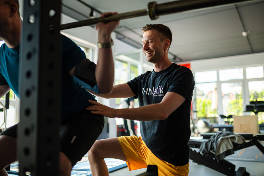 Ein Coach von x-thletik unterstützt eine trainierende Person beim Rückentraining mit Langhantel in einem modernen Studio. Die Szene zeigt die korrekte Übungsausführung und betont die Bedeutung gezielter Prävention gegen Rückenblockaden.