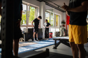 Mehrere Personen stehen in einem Trainingsraum von x-thletik, eine Trainerin beobachtet einen Athleten während einer Vorbereitungsübung. Das Bild unterstreicht die Bedeutung strukturierter Warm-up- und Cool-down-Phasen im funktionellen Training.