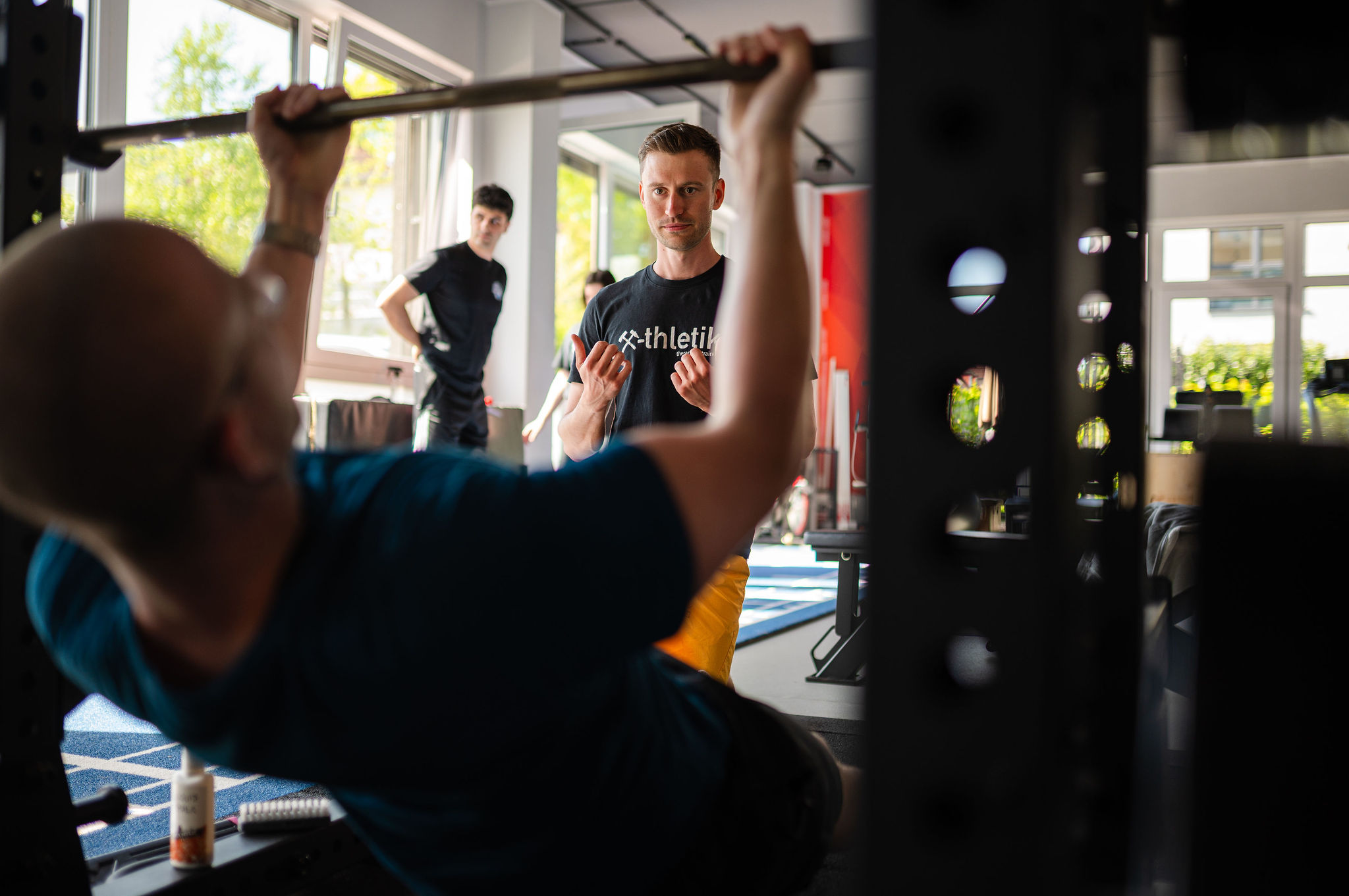 Ein x-thletik Trainer beobachtet konzentriert einen Sportler beim Langhanteltraining, während weitere Personen im Hintergrund die Szene verfolgen – Symbol für die enge Zusammenarbeit von Physiotherapie und Leistungssport.