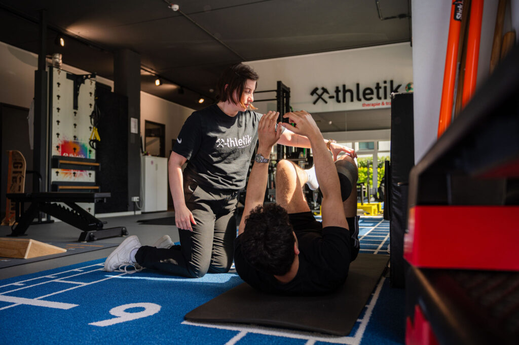 Eine x-thletik-Mitarbeiterin unterstützt eine liegende Person bei der Durchführung einer Rückenübung auf einer Trainingsmatte in einem modernen Fitnessstudio. Die Szene zeigt eine individuelle Betreuung zur gezielten Behandlung von Rückenschmerzen.