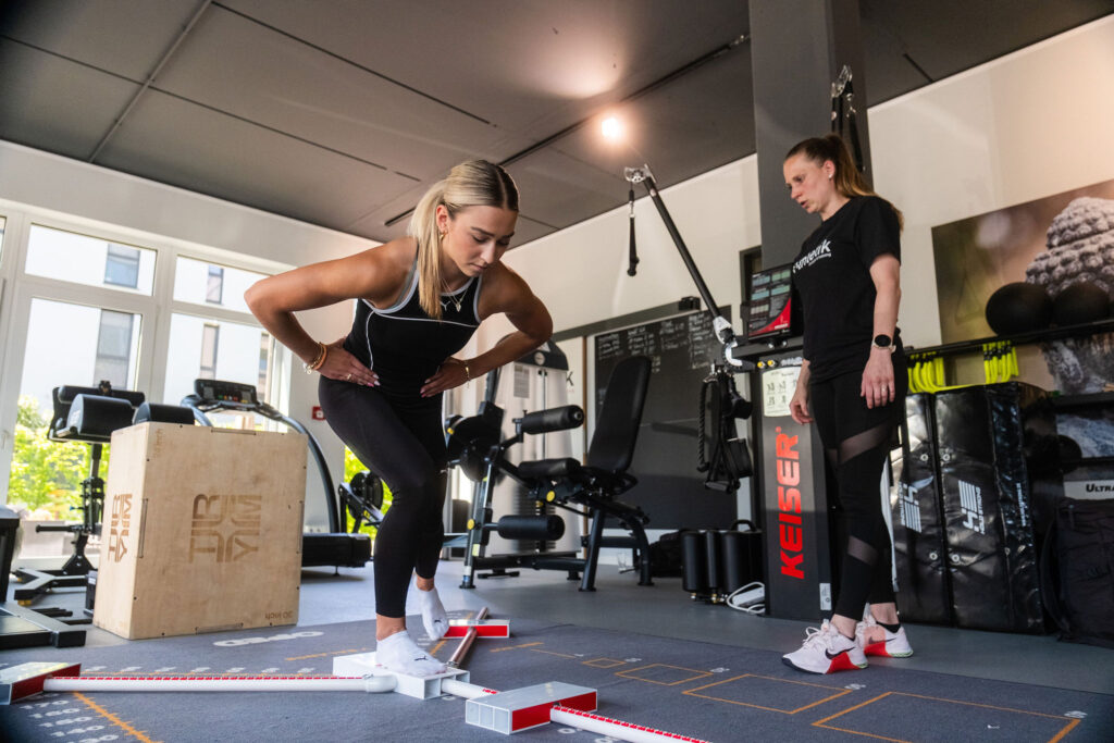 Eine Trainerin von x-thletik beobachtet konzentriert, wie eine Frau eine Gleichgewichtsübung mit einem funktionellen Testgerät ausführt. Das Bild unterstreicht die individuelle Trainingsbetreuung zur gezielten Vorbereitung auf die Marathonsaison.