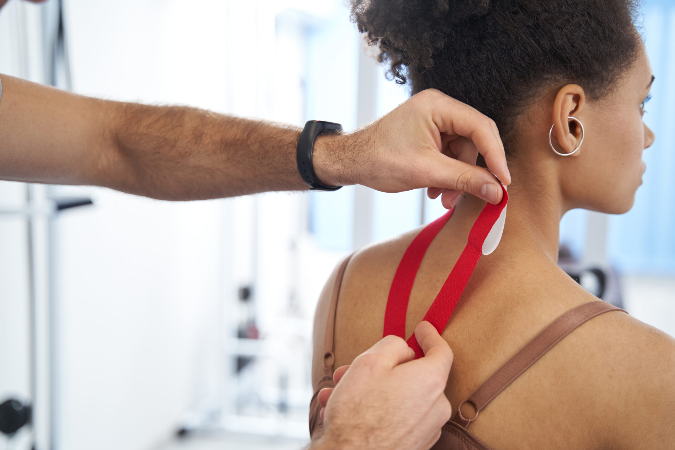 Geschultes Fachpersonal bringt rotes Kinesiotape auf dem oberen Rücken und Nacken einer jungen Frau mit lockigem Haar an. Die Tapeanlage erfolgt präzise entlang der Nackenmuskulatur, im Hintergrund ist unscharf ein Trainingsraum erkennbar.