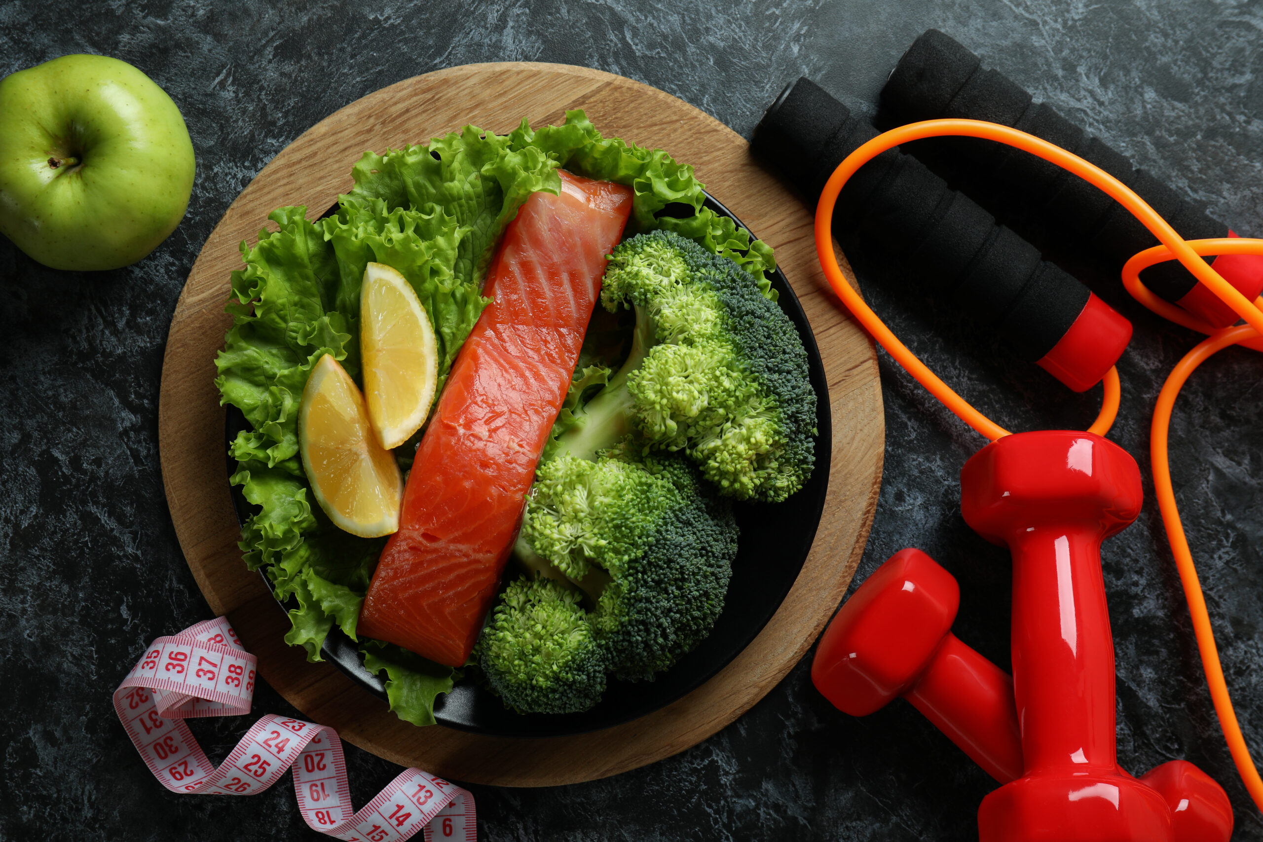 Gesunde Mahlzeit mit Lachs, Brokkoli und Zitronenspalten auf Salat, daneben Apfel, Hanteln, Springseil und Maßband – Symbol für Ernährung und Muskelregeneration.