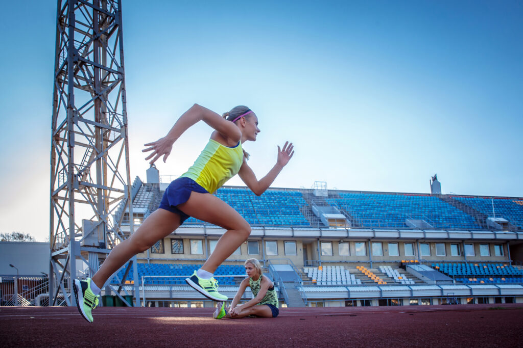 Sprinterin beim Training im Stadion, im Vordergrund in voller Bewegung, während eine Athletin im Hintergrund Dehnübungen macht – Symbol für Prävention und Physiotherapie in der Leichtathletik.