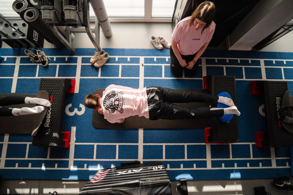 Zwei Personen beim Athletiktraining in einem Studio: Eine trainiert in Plank-Position auf einer Matte, die andere beobachtet anleitend, Fokus auf Rumpfstabilität und funktionelle Ausführung.
