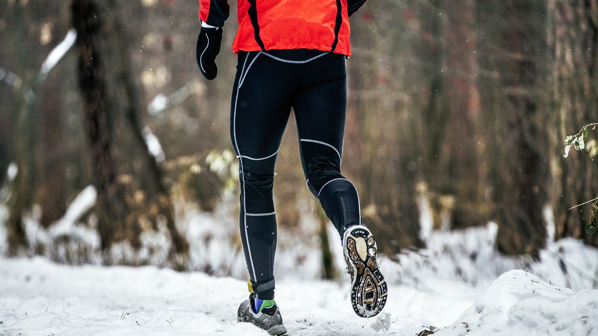 Person beim Laufen auf einem verschneiten Waldweg. Zu sehen sind die Beine von hinten, bekleidet mit schwarzer Laufhose und roter Jacke. Der Läufer trägt Trailschuhe und bewegt sich bei winterlichen Bedingungen durch eine natürliche Umgebung.