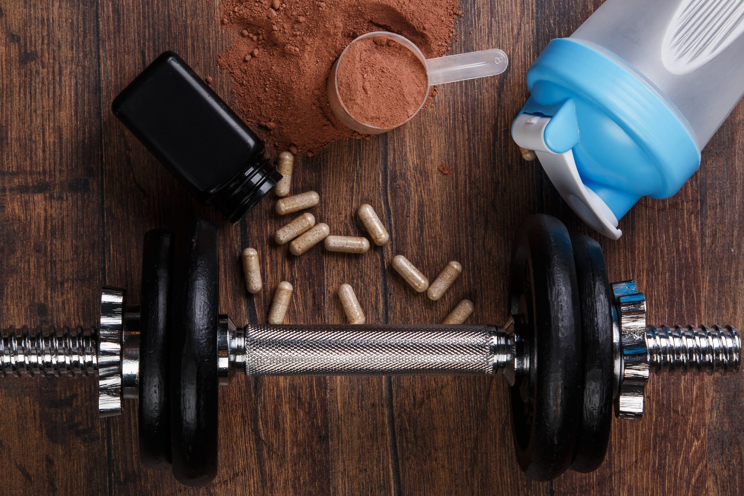 Hantel, Pulver und Kapseln liegen auf einem Holztisch. Ein Shaker, Supplement-Kapseln und Proteinpulver umgeben das Trainingsgewicht. Das Bild zeigt typische Sport-Supplements wie Kreatin, Koffein und Proteinpulver im Umfeld von Krafttraining.