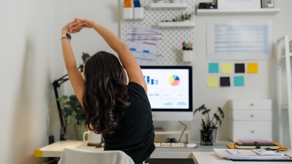 Frau dehnt Arme und Oberkörper am Schreibtisch im Homeoffice bei einer einfachen Büroübung gegen Verspannungen im Arbeitsalltag.
